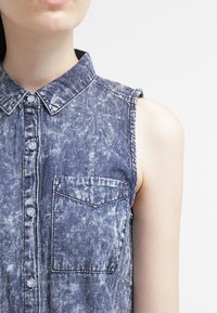 Camicetta senza maniche in denim di colore blu scuro con un lavaggio sbiadito, chiusura a bottoni sul davanti, colletto a punta e taschino sul lato sinistro.