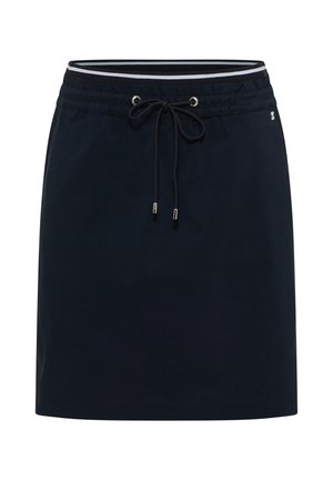 Femme portant un sweat à capuche blanc à lacets, une jupe noire à cordon et des baskets argentées, debout contre un mur beige, regardant sur le côté.