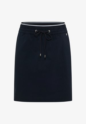 Mujer con sudadera blanca con cordones, falda negra con cordón y zapatillas plateadas, de pie contra una pared beige, mirando de lado.