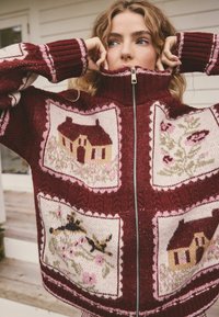 Bordeauxroter Strickpullover mit Reißverschluss, verziert mit floralen und Hausmotiven in Creme und Rosa, mit gerippten Bündchen und hohem Kragen.