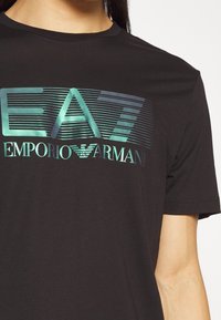 Svart t-shirt med kort ärm som har en glänsande, flerfärgad grafisk logotyp som säger "EA7 EMPORIO ARMANI" över bröstområdet.