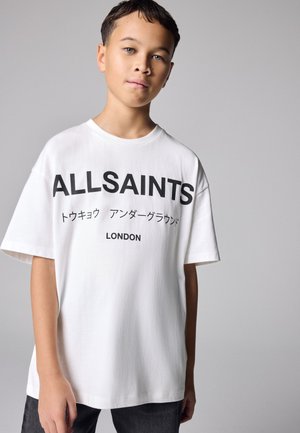 Garçon portant un t-shirt blanc AllSaints avec texte noir en japonais et en anglais, debout devant un fond gris uni.