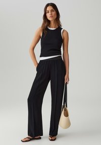 Top senza maniche nero con bordi bianchi, abbinato a pantaloni neri larghi. La modella tiene una borsa di paglia e indossa infradito nere. Outfit semplice e casual.