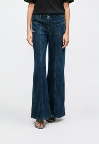 Sötétkék, széles szárú farmert viselő személy, elülső tapaszzsebekkel, valamint fekete, vékony pántos nyitott orrú szandált visel, egyszerű háttér előtt.