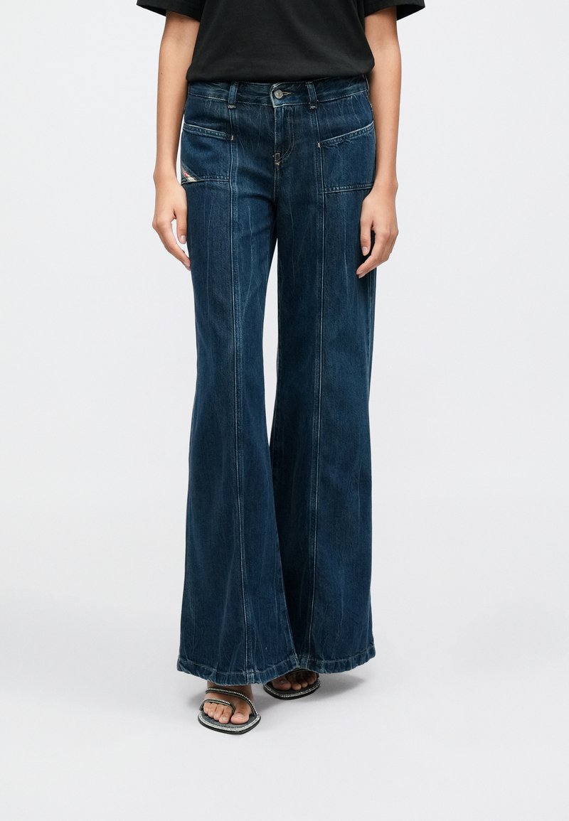 Sötétkék, széles szárú farmert viselő személy, elülső tapaszzsebekkel, valamint fekete, vékony pántos nyitott orrú szandált visel, egyszerű háttér előtt.