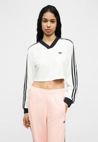 Γυναίκα με μακριά σκούρα μαλλιά φορά λευκό και μαύρο κοντό φούτερ και ανοιχτό ροζ παντελόνι φόρμας, στέκεται μπροστά σε απλό ανοιχτό φόντο.