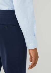Camicia a quadre blu chiaro con bottoni bianchi, abbinata a pantaloni navy con tasca posteriore visibile e etichetta con il marchio.