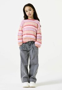 Strickpullover mit pink, weiß und gelb gestreift, kombiniert mit hochtaillierten grauen Denimhosen. Das Model trägt weiße Sneaker. Einfacher Hintergrund.