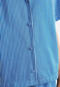 Kortärmad blå skjorta med vertikala vita ränder, med knäppningar och rak nederkant. Mjuk, lätt tygtextur.