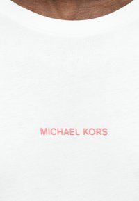 Λευκό πουκάμισο από ύφασμα με την επιγραφή "MICHAEL KORS" σε ροζ γράμματα, κεντραρισμένη κάτω από το γιακά σε σκουρόχρωμο δέρμα.