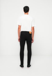 Hombre de pie, de espaldas, vistiendo una camiseta blanca lisa, jeans negros ajustados y zapatillas negras con suelas blancas, sobre un fondo blanco.