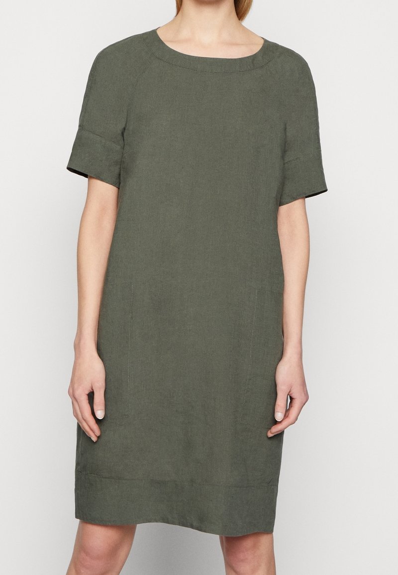 Femme portant une robe ample à manches courtes vert olive avec un col rond, debout devant un fond clair uni.