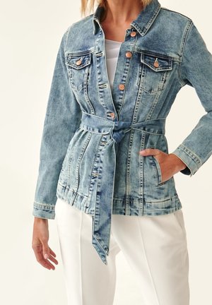 Denimjakke med knapper foran, brystlommer og bundet bælte, båret over hvid top og hvide bukser, modellens højre hånd i lommen.