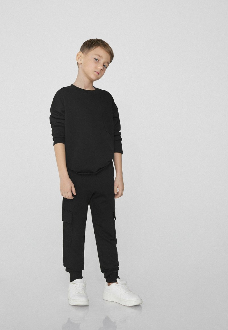 Černá mikina s přední kapsou, spárovaná s černými cargo kalhotami se bočními kapsami a žebrovanými manžetami, doplněná bílými teniskami.