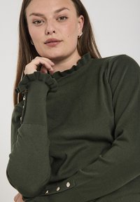 Donkergroene gebreide trui met een gerimpelde halslijn en manchetten, versierd met knopen aan de zijkant. Glad textuur met een comfortabele pasvorm.