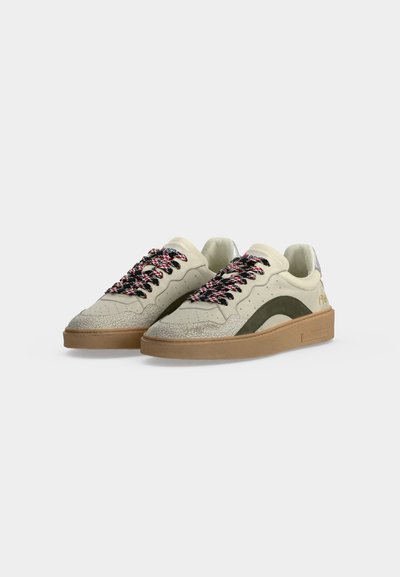 Paire de baskets basses écrues avec semelles marron, lacets noirs et rouges à motifs, et accents latéraux vert foncé sur un fond uni.
