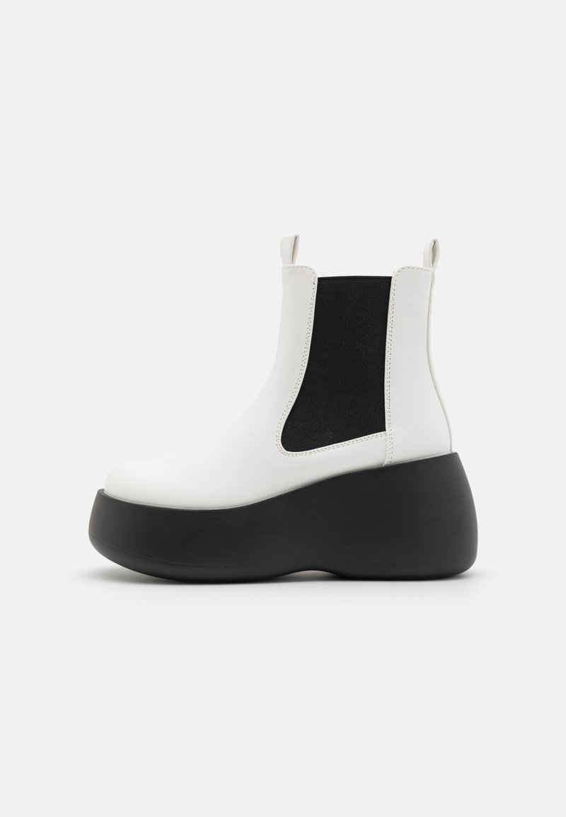 Bottine blanche à cheville avec un panneau latéral élastique noir et une semelle plateforme noire épaisse. Tige en cuir lisse avec des languettes pour un enfilage facile.