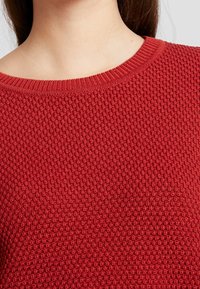 Maglione rosso lavorato a maglia con un motivo testurizzato, dotato di un ampio scollo rotondo e polsini a coste. La vista ravvicinata mostra i dettagli della cucitura.