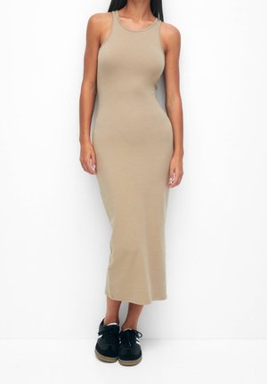 Robe maxi beige sans manches en tissu extensible, design ajusté, texture lisse, avec un col rond et une fente sur le côté, associée à des baskets noires.