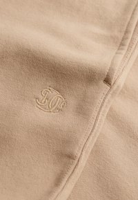 Beżowy bawełniany strój o miękkiej fakturze, z haftowanym logo. Widoczne są detale szwów, podkreślające gładki design.