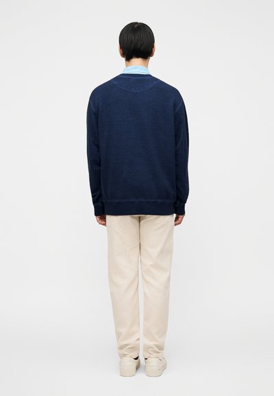 Σκούρο μπλε φούτερ με υφή και ριμπ μανσέτες, σε συνδυασμό με μπεζ παντελόνι και ανοιχτόχρωμα παπούτσια. Μόνο πίσω όψη.