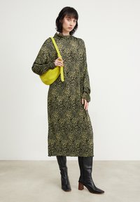 Robe longue à imprimé léopard noir et vert avec un col haut ; associée à des bottes à talons noires jusqu'aux genoux et un petit sac jaune.