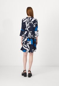 Vestido floral azul-marinho com padrões em branco e azul, mangas em sino, design em camadas, comprimento até ao joelho, combinado com sandálias pretas robustas.
