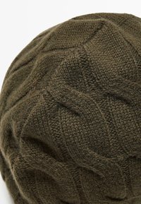 Béret en tricot vert olive foncé avec un motif en losange surélevé et une couture texturée, vu du dessus sur un fond blanc.