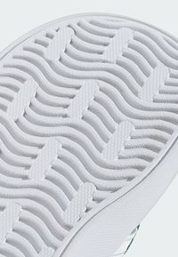 Bílá gumová podrážka boty s texturovaným vlnitým vzorem, obsahující vyvýšené sekce pro lepší grip a ploché oblasti pro zvýšenou stabilitu.