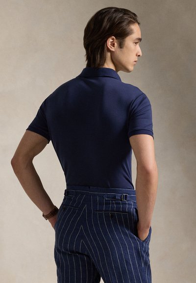 Mann trägt dunkelblaues Poloshirt und blaue Nadelstreifenhose, steht seitlich mit Händen in den Taschen vor beigem Hintergrund.