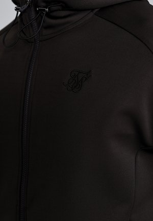 Black zip-up jacket made of smooth fabric, featuring a raised logo on the chest, high collar, and adjustable drawstring.