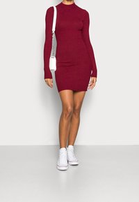 Vestito bordeaux a coste, con maniche lunghe, colletto alto e linea aderente, abbinato a sneakers alte bianche e una piccola borsa bianca.