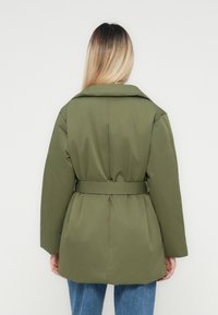 Blond person som bär en olivgrön kappa med bälte och blå jeans, sedd bakifrån mot en enkel ljus bakgrund.