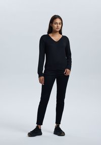 Zwarte lange mouwtop met een V-hals, gecombineerd met zwarte slim-fit broeken en zwarte sneakers met gouden accenten. Simpel, gestroomlijnd ontwerp.