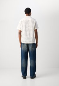 Witte shirt met korte mouwen en een textuur van golven, gecombineerd met blauwe losvallende jeans. Model is van achteren gezien en draagt zwarte schoenen.