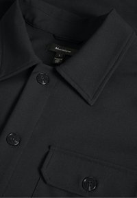 Chemise noire à col avec boutons visibles et poche sur le devant, montrant une étiquette de marque "Matinique" et une étiquette de taille à l'intérieur du col.