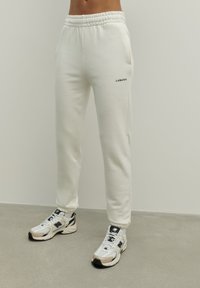 Białe bawełniane spodnie dresowe o luźnym kroju, z elastycznym paskiem w talii, bocznymi kieszeniami i napisem "LAMANIA" na lewej nodze. W komplecie z sportowymi butami.