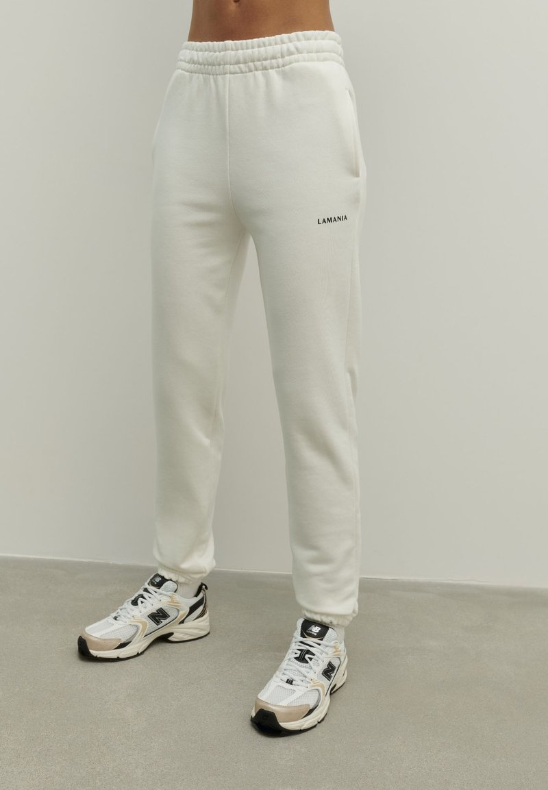 Białe bawełniane spodnie dresowe o luźnym kroju, z elastycznym paskiem w talii, bocznymi kieszeniami i napisem "LAMANIA" na lewej nodze. W komplecie z sportowymi butami.