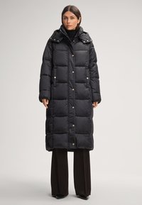 Cappotto puffer nero con un design lungo e squadrato, dotato di cappuccio, bottoni a pressione e sezioni trapuntate orizzontali. Abbinato a pantaloni scuri.