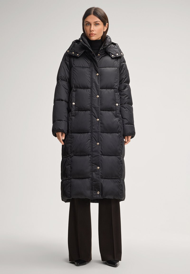 Cappotto puffer nero con un design lungo e squadrato, dotato di cappuccio, bottoni a pressione e sezioni trapuntate orizzontali. Abbinato a pantaloni scuri.