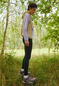 Chaqueta ligera beige y lavanda con capucha y un bolsillo frontal, combinada con leggins negros y zapatillas de correr grises en un entorno natural.