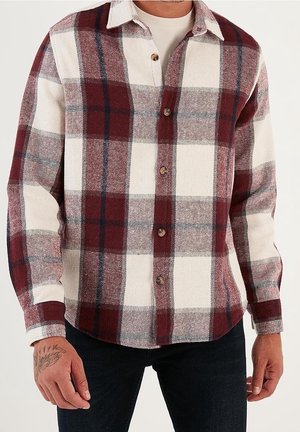 Mann trägt ein langärmliges Hemd mit großem rot-weiß-navy Karomuster über einem weißen Unterhemd.