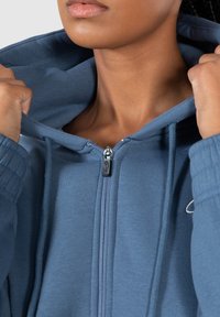 Sudadera azul con cremallera, de textura suave, capucha ajustable y puños acanalados. Presenta una cremallera metálica y un discreto detalle del logo en el pecho.