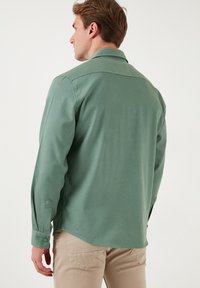 Cămașă lungă verde cu mâneci lungi, featuring un guler cu nasturi, buzunare laterale și jambe ajustabile, asortată cu pantaloni bej. Textură de țesătură netedă.