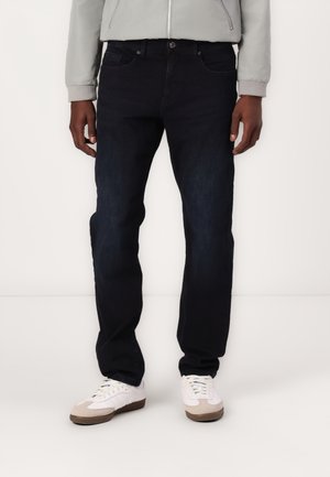 Mand iført sorte slim-fit jeans, hvide sneakers med beige accenter og en lysegrå lynlåsjakke, stående mod en ensfarvet baggrund.