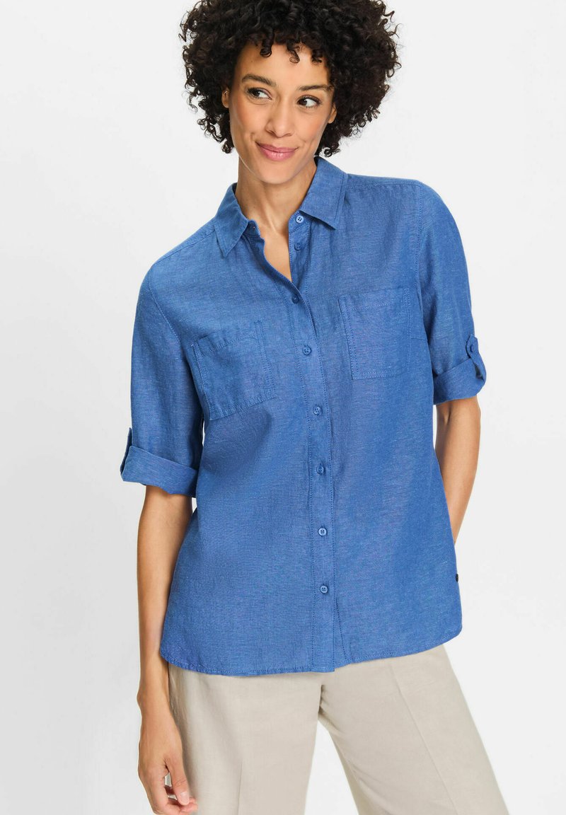 Blue button-down shirt in lightweight fabric, featuring two chest pockets, rolled sleeves, and a curved hem. Soft texture with a slight sheen.