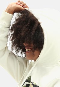 Biała bluza z kapturem z kwiatowym wzorem, strukturalna tkanina, długie rękawy i regulowany kaptur, przedstawiona jako zakładana na kręcone włosy.