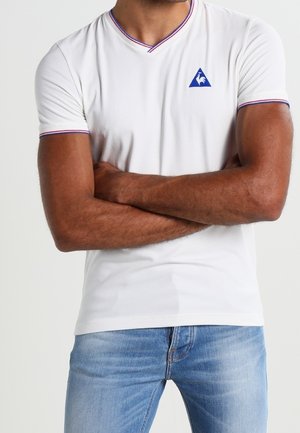 Weißes T-Shirt mit kurzen Ärmeln und V-Ausschnitt, akzentuiert mit lila Besatz und blauem Logo auf der linken Brust. Weicher Stoff.