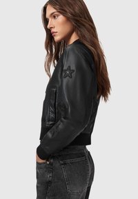 Chaqueta bomber de cuero negro con estrellas y corazones bordados en las mangas, puños y cintura acanalados, con cierre de cremallera en la parte delantera y bolsillos laterales.
