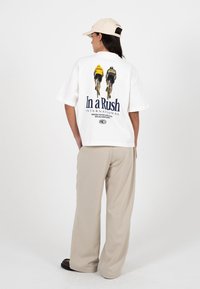 Femme portant un t-shirt blanc avec des cyclistes et le texte « In a Rush International » au dos, pantalon beige, casquette crème, debout devant un fond blanc.